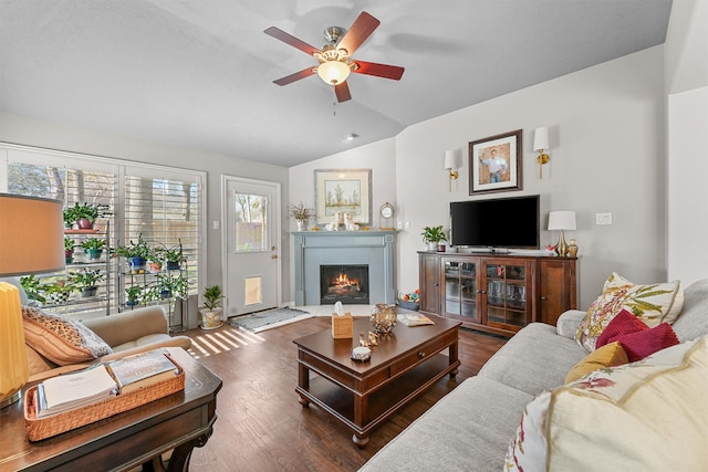 living area featuring vaulted ceiling, ceiling fan, a warm lit fireplace, and wood finished floors