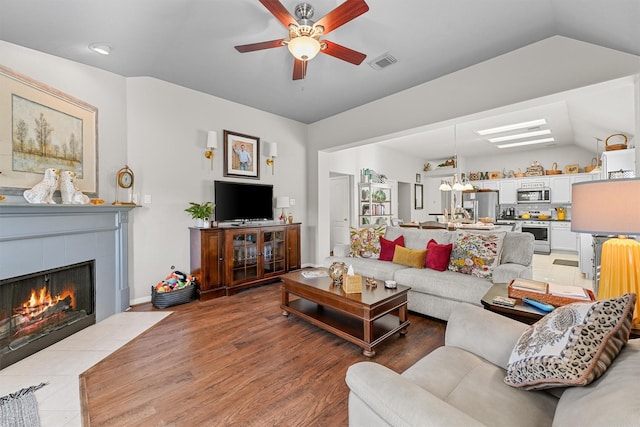 living room featuring visible vents, light wood-style floors, vaulted ceiling, ceiling fan, and a tile fireplace