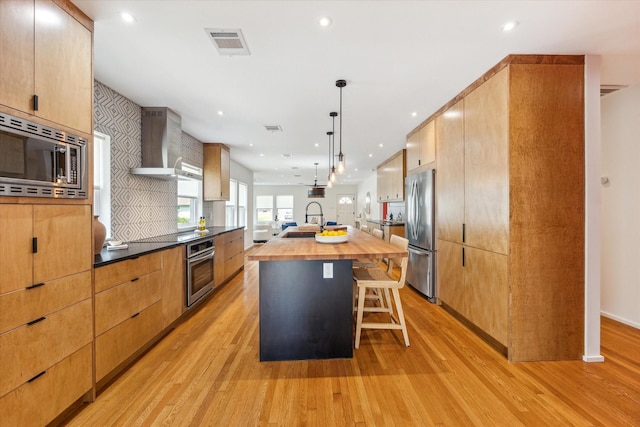 kitchen with wall chimney exhaust hood, stainless steel appliances, a large island with sink, pendant lighting, and decorative backsplash