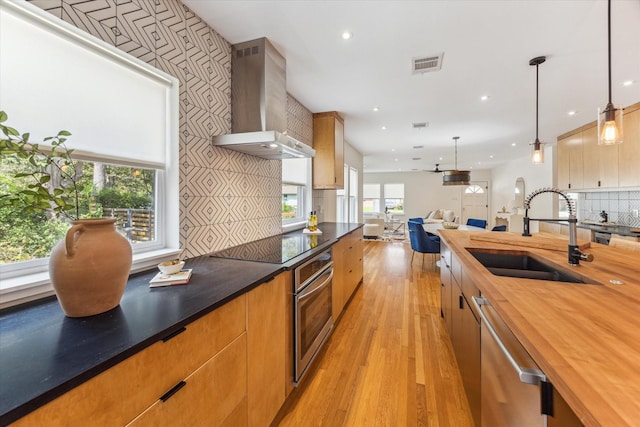 kitchen featuring sink, wall chimney exhaust hood, hanging light fixtures, appliances with stainless steel finishes, and light wood-type flooring