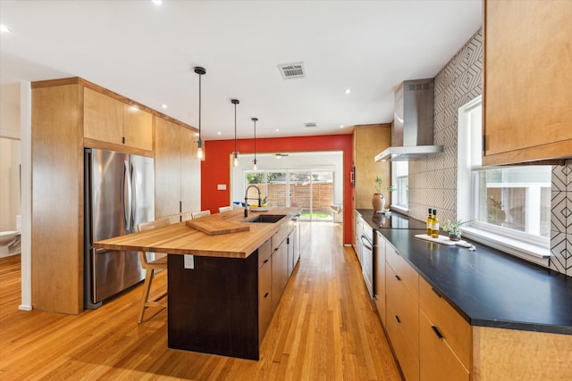 kitchen featuring pendant lighting, a kitchen island with sink, sink, wall chimney exhaust hood, and stainless steel refrigerator