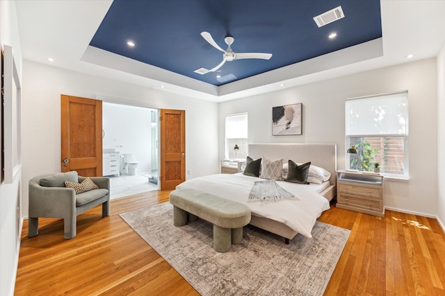 bedroom featuring a tray ceiling, ensuite bath, ceiling fan, and light wood-type flooring