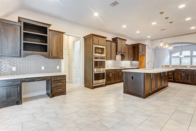 kitchen with dark brown cabinetry, tasteful backsplash, an island with sink, pendant lighting, and appliances with stainless steel finishes