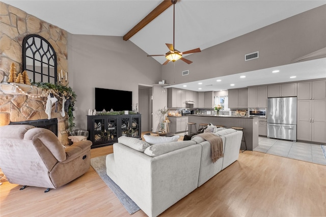 living room with beam ceiling, ceiling fan, a stone fireplace, high vaulted ceiling, and light wood-type flooring