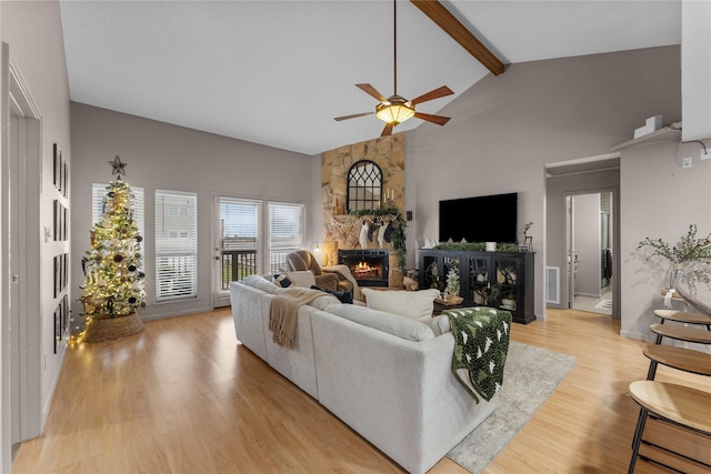 living room with beam ceiling, ceiling fan, a stone fireplace, high vaulted ceiling, and light wood-type flooring