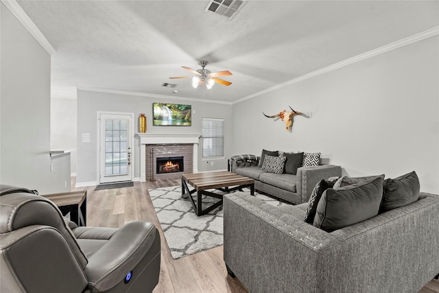 living room featuring a brick fireplace, ornamental molding, a textured ceiling, ceiling fan, and light hardwood / wood-style flooring