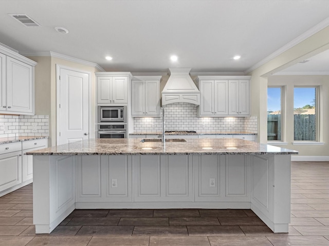 kitchen featuring light stone counters, a center island with sink, appliances with stainless steel finishes, and custom exhaust hood