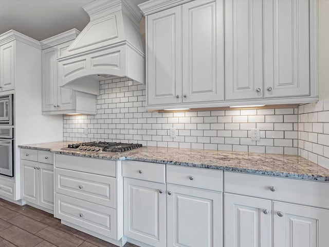 kitchen with stainless steel appliances, light stone counters, backsplash, white cabinets, and custom exhaust hood