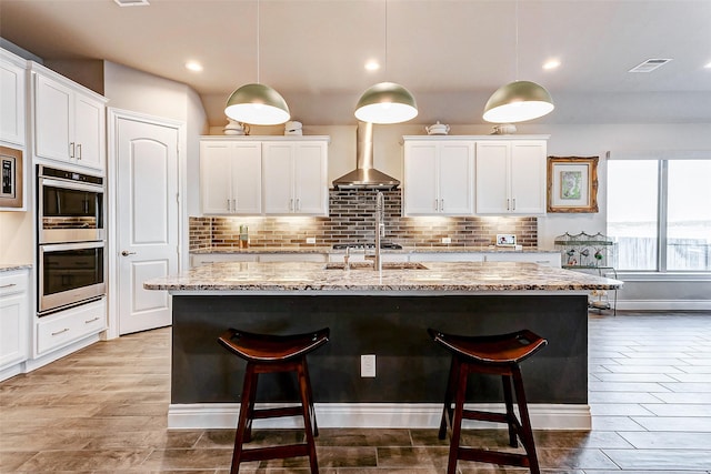 kitchen featuring hanging light fixtures, an island with sink, and wall chimney exhaust hood