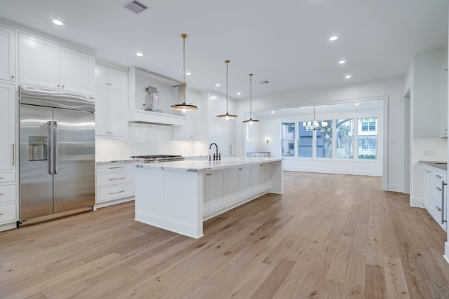 kitchen featuring hanging light fixtures, stainless steel built in fridge, white cabinets, a center island with sink, and light wood-type flooring