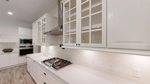 kitchen with appliances with stainless steel finishes, white cabinets, decorative backsplash, light hardwood / wood-style floors, and wall chimney exhaust hood