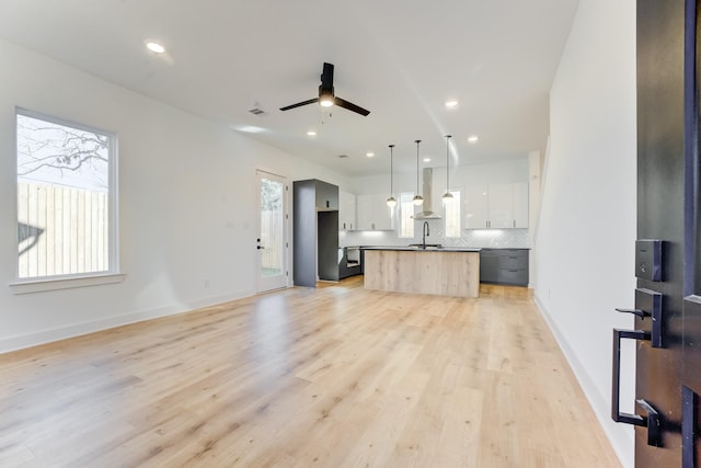 kitchen featuring decorative backsplash, a kitchen island with sink, wall chimney range hood, decorative light fixtures, and white cabinets