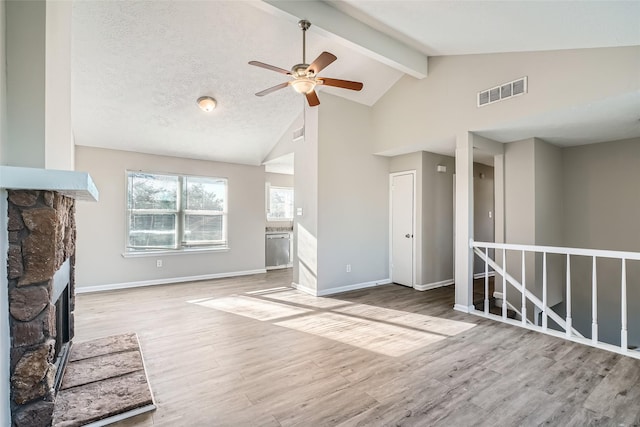 unfurnished living room with a textured ceiling, ceiling fan, lofted ceiling with beams, light hardwood / wood-style floors, and a stone fireplace