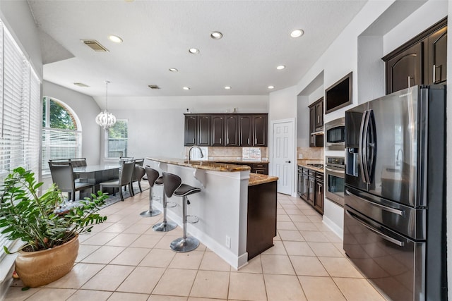 kitchen featuring backsplash, a center island with sink, hanging light fixtures, appliances with stainless steel finishes, and dark brown cabinetry