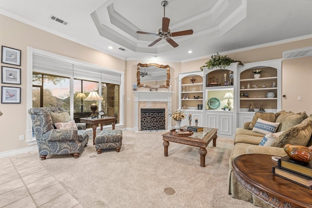 living room with a high end fireplace, ornamental molding, built in shelves, a tray ceiling, and ceiling fan