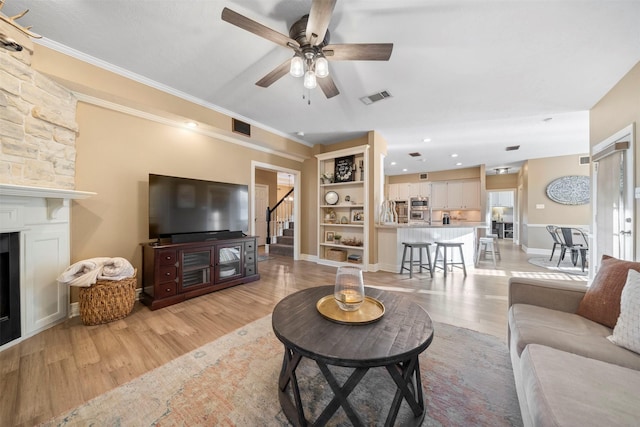 living room featuring crown molding, a stone fireplace, light hardwood / wood-style floors, and ceiling fan