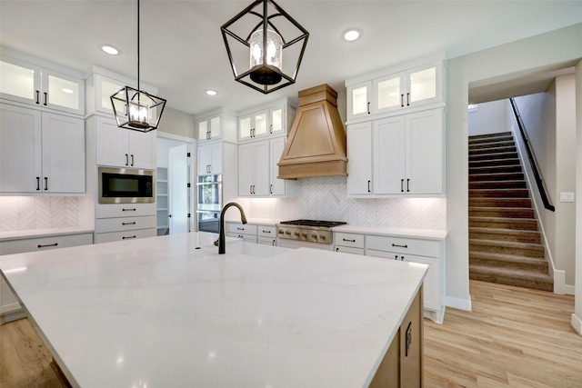 kitchen with decorative light fixtures, white cabinets, premium range hood, a kitchen island with sink, and appliances with stainless steel finishes