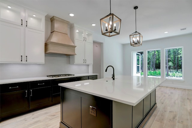 kitchen with premium range hood, a center island with sink, white cabinets, hanging light fixtures, and light hardwood / wood-style flooring