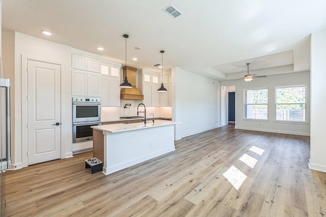 kitchen with a center island with sink, premium range hood, white cabinetry, stainless steel double oven, and a raised ceiling