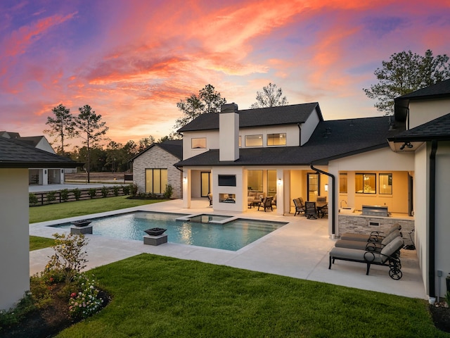 back house at dusk featuring a yard, exterior kitchen, a swimming pool with hot tub, a fireplace, and a patio