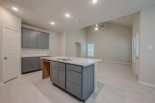 kitchen featuring light stone counters, ceiling fan, a kitchen island with sink, sink, and gray cabinets