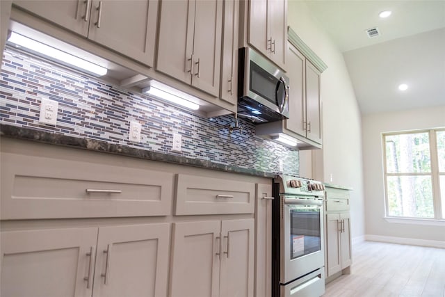 kitchen featuring stainless steel appliances, backsplash, dark stone counters, lofted ceiling, and light wood-type flooring