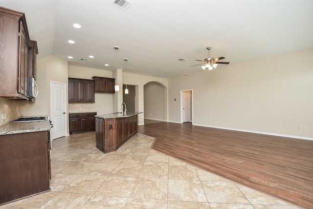 kitchen featuring dark brown cabinetry, light stone countertops, ceiling fan, hanging light fixtures, and a center island with sink