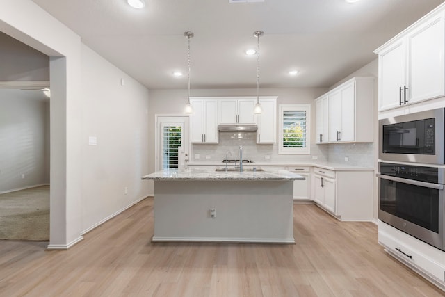 kitchen featuring pendant lighting, oven, an island with sink, white cabinetry, and black microwave