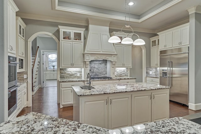 kitchen featuring a kitchen island with sink, a raised ceiling, hanging light fixtures, decorative backsplash, and stainless steel appliances