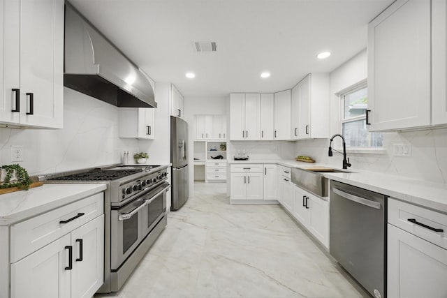 kitchen featuring light stone counters, wall chimney exhaust hood, stainless steel appliances, sink, and white cabinets