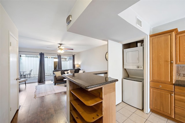 kitchen featuring ceiling fan, kitchen peninsula, dark stone counters, stacked washer / drying machine, and light tile patterned floors