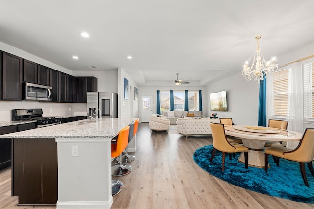 kitchen featuring a tray ceiling, hanging light fixtures, a kitchen island with sink, stainless steel appliances, and ceiling fan with notable chandelier