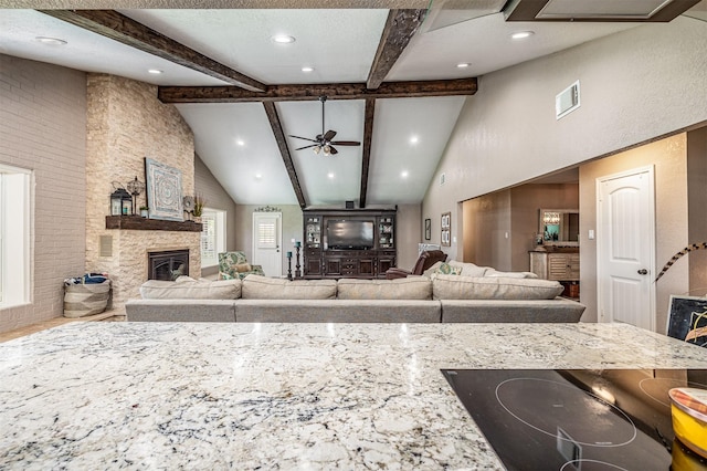 kitchen featuring light stone counters, ceiling fan, beam ceiling, high vaulted ceiling, and a stone fireplace