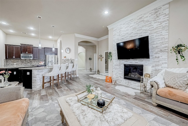 living room featuring arched walkways, ornamental molding, a stone fireplace, light wood-type flooring, and recessed lighting