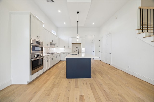 kitchen featuring a kitchen island with sink, gas stovetop, stainless steel double oven, decorative light fixtures, and white cabinetry