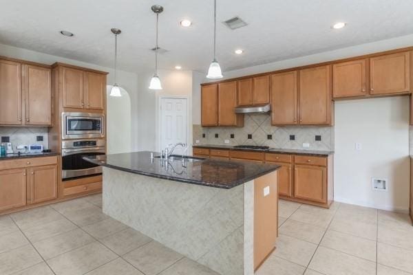 kitchen with appliances with stainless steel finishes, dark stone counters, a kitchen island with sink, sink, and hanging light fixtures