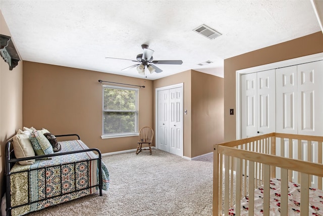 carpeted bedroom with a textured ceiling, two closets, ceiling fan, and a crib