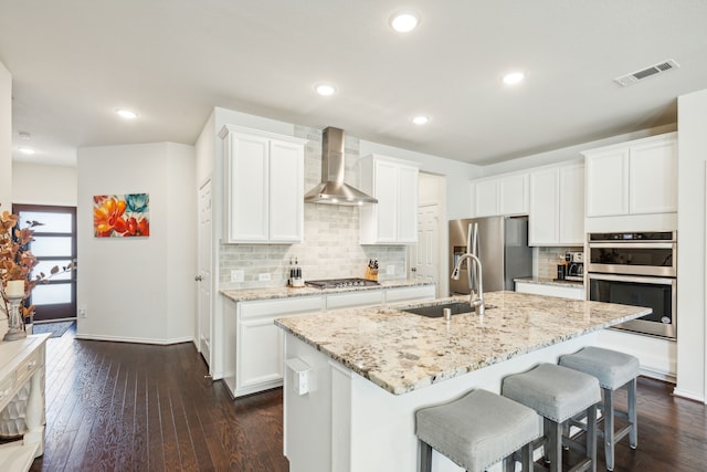 kitchen with a kitchen island with sink, white cabinets, wall chimney range hood, and appliances with stainless steel finishes