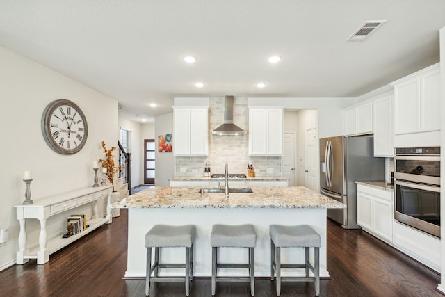 kitchen featuring stainless steel appliances, wall chimney range hood, light stone counters, a kitchen island with sink, and white cabinets