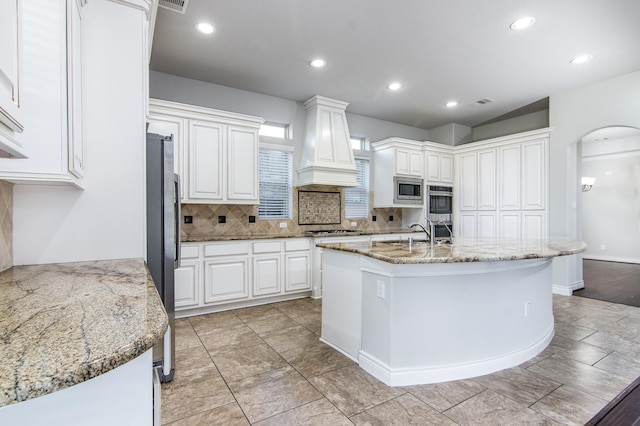 kitchen featuring custom exhaust hood, a kitchen island with sink, light stone countertops, appliances with stainless steel finishes, and white cabinetry