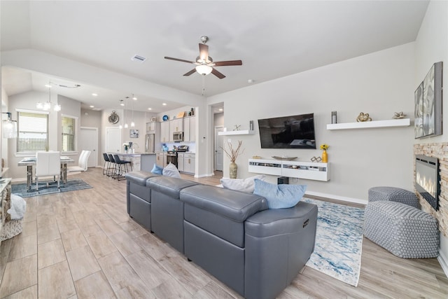 living room featuring ceiling fan with notable chandelier, vaulted ceiling, light hardwood / wood-style flooring, and a stone fireplace