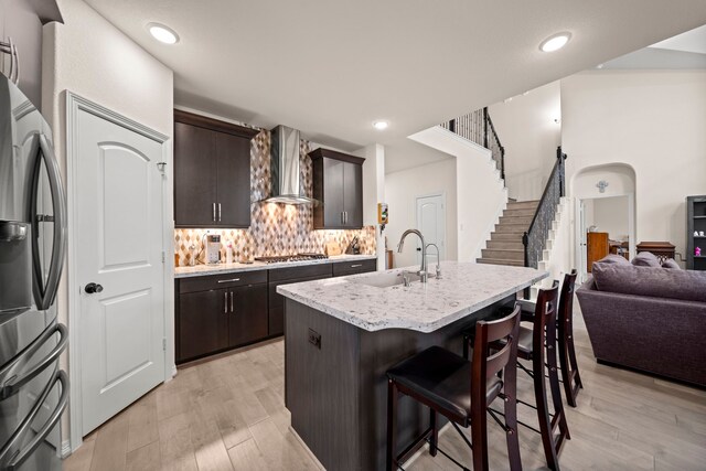 kitchen with appliances with stainless steel finishes, wall chimney exhaust hood, dark brown cabinets, sink, and a center island with sink