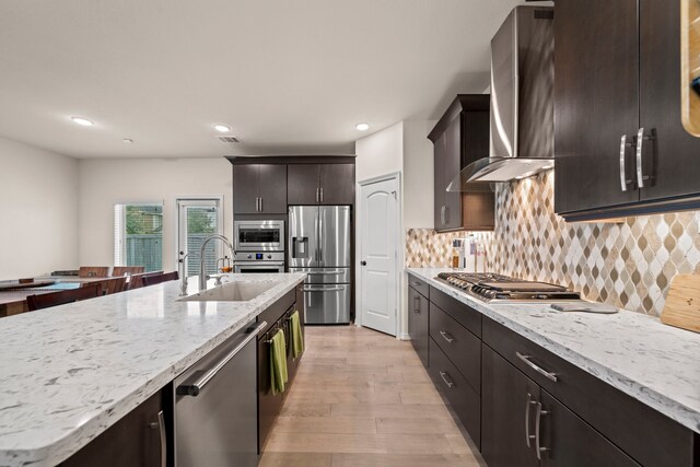 kitchen featuring wall chimney exhaust hood, light wood-type flooring, appliances with stainless steel finishes, tasteful backsplash, and dark brown cabinetry