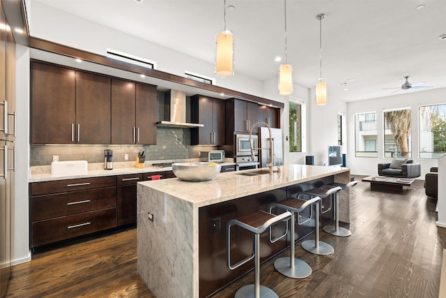 kitchen with ceiling fan, pendant lighting, decorative backsplash, an island with sink, and wall chimney exhaust hood