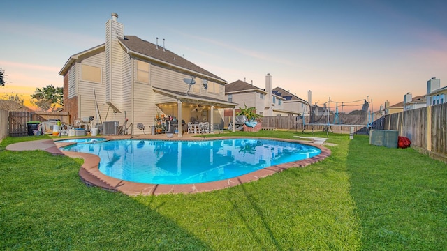 pool at dusk with a lawn, a diving board, cooling unit, and a trampoline