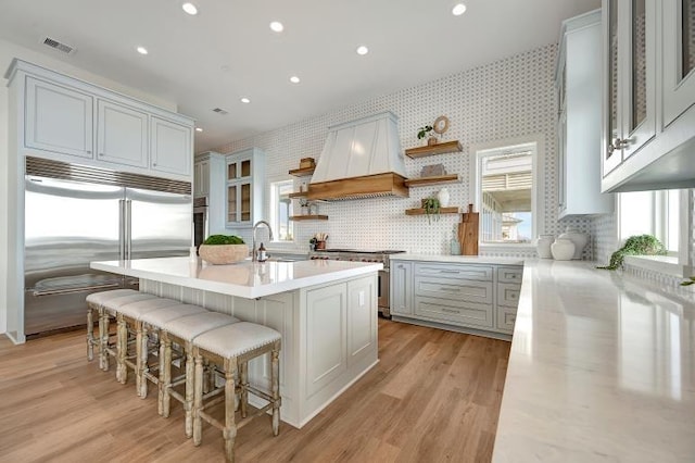 kitchen featuring a kitchen bar, custom range hood, a kitchen island with sink, light hardwood / wood-style flooring, and high quality appliances