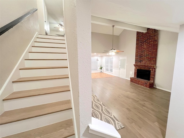 stairs featuring ceiling fan, wood-type flooring, lofted ceiling with beams, and a brick fireplace