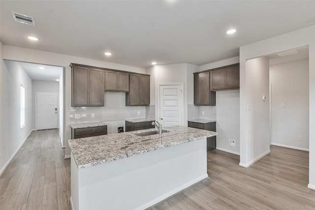 kitchen with sink, light stone countertops, light wood-type flooring, an island with sink, and tasteful backsplash