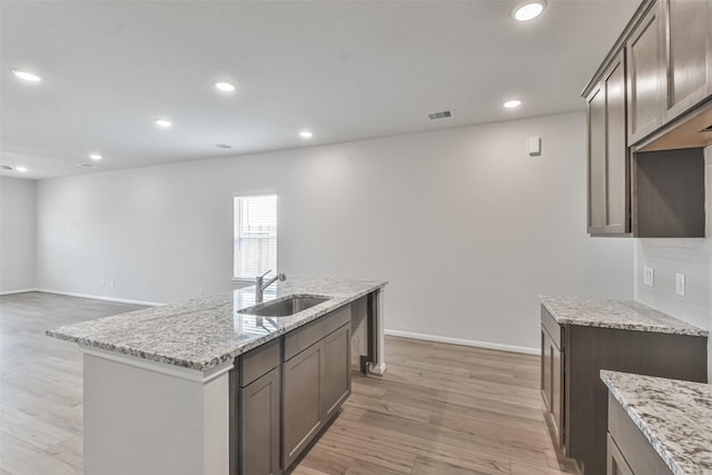 kitchen with a center island with sink, sink, light wood-type flooring, dark brown cabinets, and light stone counters