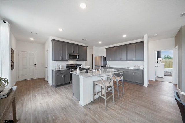 kitchen with appliances with stainless steel finishes, a kitchen breakfast bar, light stone counters, a kitchen island with sink, and light hardwood / wood-style flooring